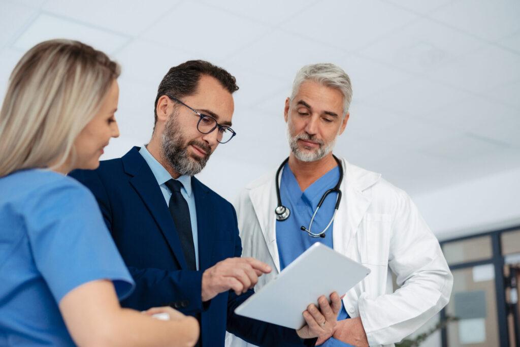 Healthcare IT consultant discussing technology solutions with medical staff in a hospital hallway, reviewing digital tools to improve efficiency and reduce vendor costs.